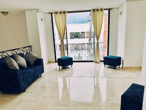 A spacious room with a large window, a sofa, two ottomans, and a metal-framed bed. - Maljut House (Cali)