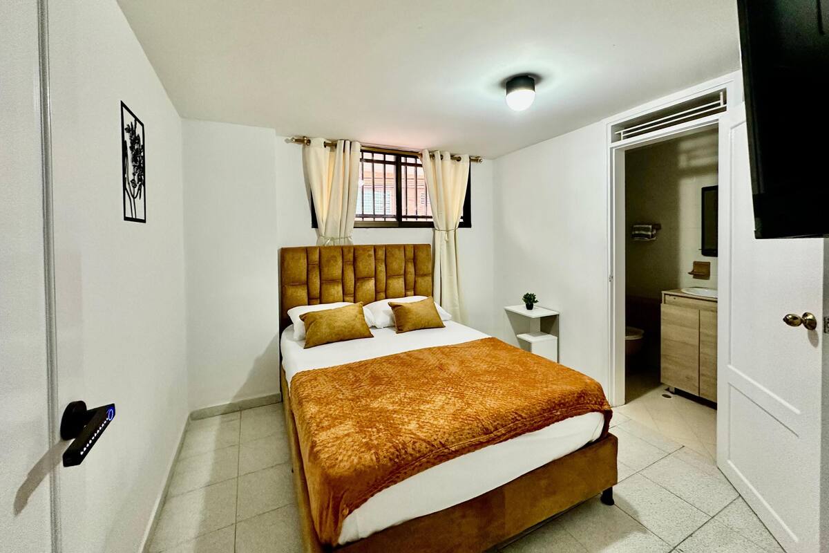 Un dormitorio con cama, mesita de noche, cortinas en la ventana y una puerta que da a otra habitación.