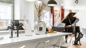 Een moderne keuken met een wit aanrecht, hangende pendantlampen, een vaas met gedroogde bloemen en een piano op de achtergrond.