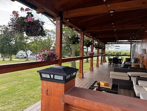 Une terrasse extérieure couverte, agrémentée de suspensions florales, de mobilier d’extérieur et offrant une vue sur un espace verdoyant avec des arbres et une maison en arrière-plan.