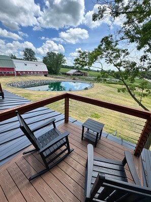 A wooden deck with outdoor furniture, a pond, and a view of a red barn and a house in the background. - Vermont Luxury Farmhouse | 5 Suites • Pond • Game Room (Milton)