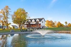 A two-story house with a large front porch, a pond with a fountain, and autumn-colored trees. - Vermont Luxury Farmhouse | 5 Suites • Pond • Game Room (Milton)