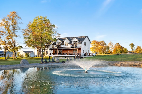 Une maison à deux étages avec une grande véranda à l’avant, un étang avec une fontaine et des arbres aux couleurs automnales.