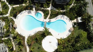 Eine Vogelperspektive zeigt ein Resort mit zwei großen Schwimmbecken, umgeben von Palmen und Liegestühlen.