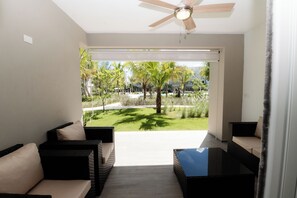 Ein modernes Wohnzimmer mit Deckenventilator, einem schwarzen Couchtisch und zwei Sofas mit beigen Kissen. Ein großes Fenster bietet Blick auf einen gepflegten Garten mit Palmen und einem Weg.