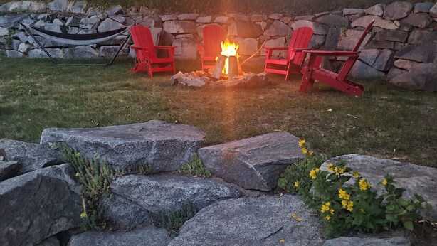 A stone fire pit with red chairs around it, set in a grassy area with large rocks and yellow flowers in the foreground. - Amazingly convenient central location in Rossland with everything you need! (Rossland)
