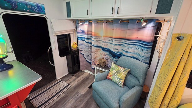 A compact living space with a kitchenette, a blue armchair, a decorative curtain, and a small table with a lamp.