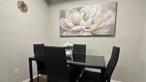 Un coin repas avec une table rectangulaire et quatre chaises, un tableau représentant des fleurs accroché au mur, et un objet décoratif en forme de soleil également fixé au mur.