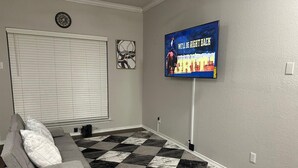 Un salon avec un canapé gris, un sol à carreaux, un téléviseur fixé au mur et une fenêtre avec des stores.