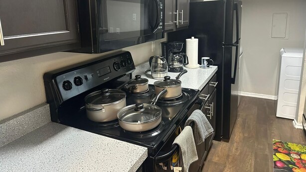 Une cuisine avec des meubles de couleur sombre, un micro-ondes noir, une cuisinière avec des casseroles, une machine à café et un réfrigérateur.