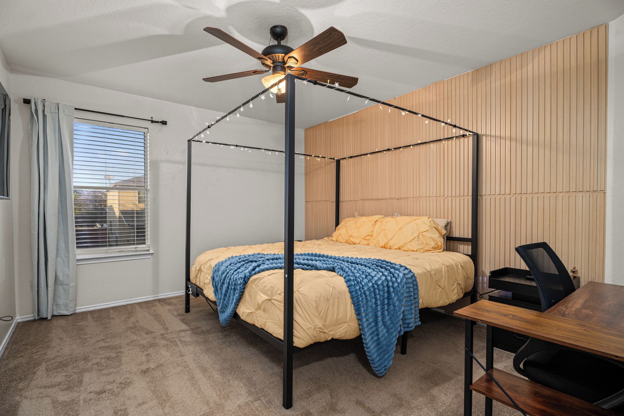 Un lit à baldaquin avec un ventilateur de plafond, un panneau mural en bois, une fenêtre avec des stores et un bureau avec une chaise.