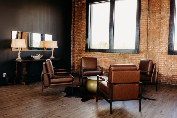 Une pièce avec un mur en briques, une grande fenêtre, des fauteuils en cuir, une table ronde et un miroir agrémenté de lampes.