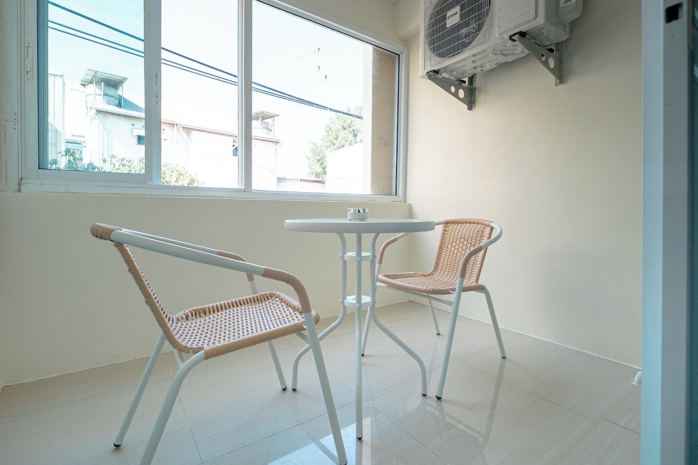 Ein Zimmer mit zwei Korbstühlen und einem kleinen Tisch, ein Fenster mit Blick auf Gebäude und eine an der Wand befestigte Klimaanlage.