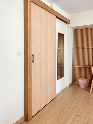 A room with a wooden sliding door, a small wooden table, a pink trash bin, and a white wall with a light switch. - The Nook Hotel (Chiang Mai)