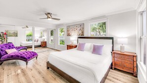 Ein Schlafzimmer mit einem großen Bett, einer violetten Liege, einem Holzbeistelltisch und einem Deckenventilator.