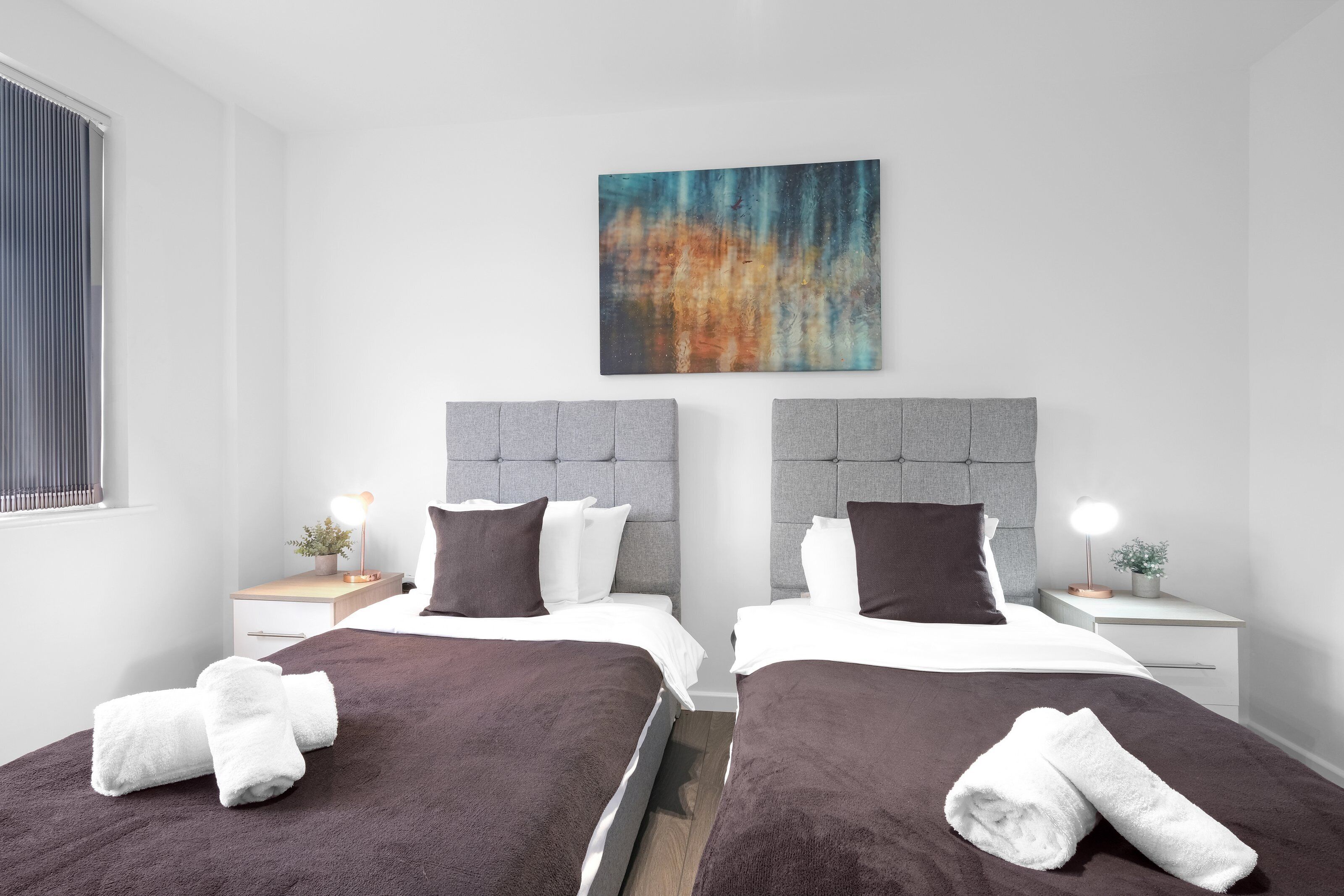 Ein Hotelzimmer mit zwei Betten, einem modernen Kopfteil, Nachttischen mit Lampen und einem großen, abstrakten Gemälde an der Wand.