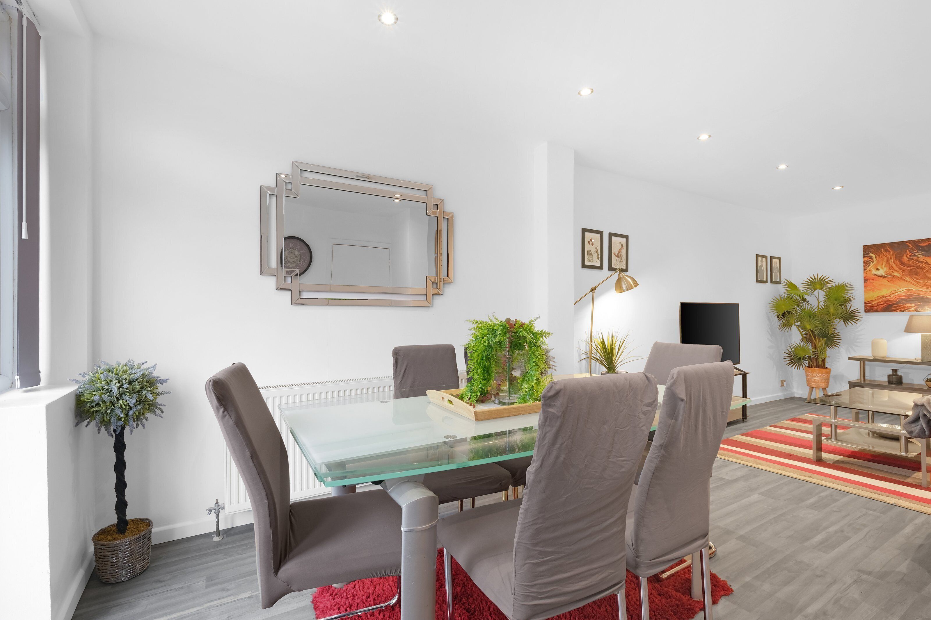 A modern dining area with a glass table, grey chairs, a red rug, and a potted plant.