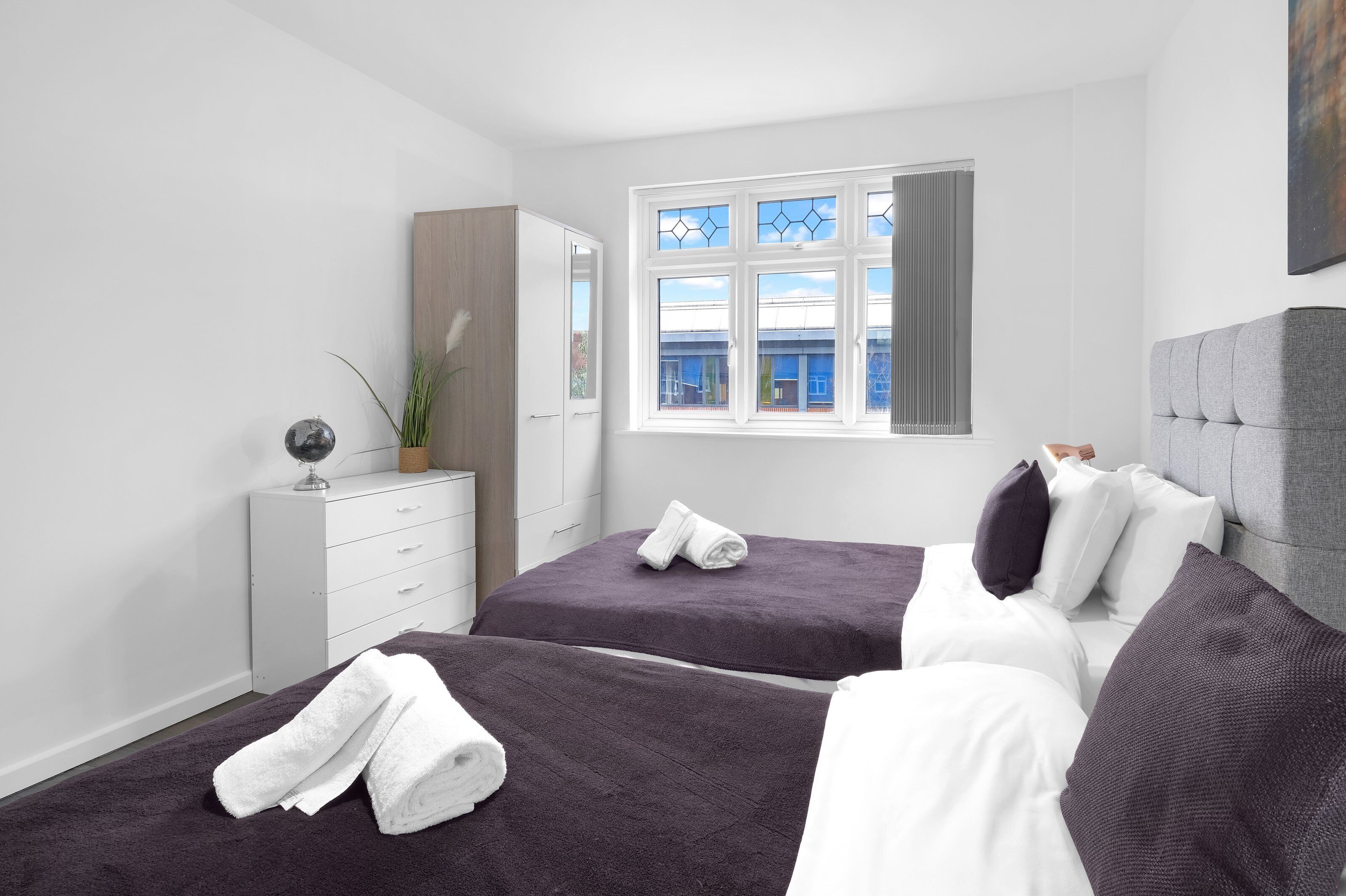Habitación de hotel con dos camas, un aparador, una ventana con vistas y un cabecero gris.