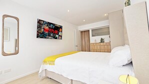 Un dormitorio con cama, espejo, televisor de pared mostrando Netflix, cómoda y ventana.
