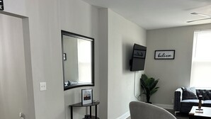 Ein modernes Wohnzimmer mit grauem Sofa, weißem Pouf, Wand-Fernseher und einem Bilderrahmen mit dem Wort 'believe'.