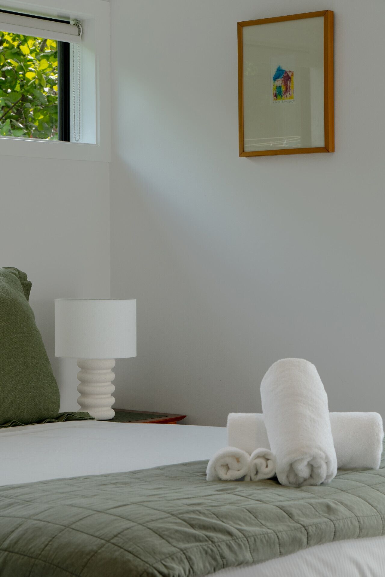 Een hotelkamer met een bed, een groen kussen, een witte lamp, een ingelijst schilderij aan de muur, en een raam met uitzicht op bomen.