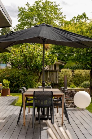 Une terrasse extérieure avec une table et des chaises, protégée par un grand parasol noir, entourée de verdure et bordée d’une terrasse en bois.