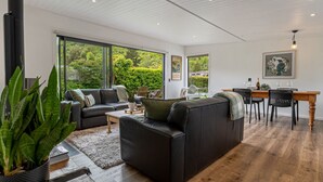 Ein modernes Wohnzimmer mit großem Fenster, einem Essbereich mit Holztisch und -stühlen sowie einem schwarzen Ledersofa.