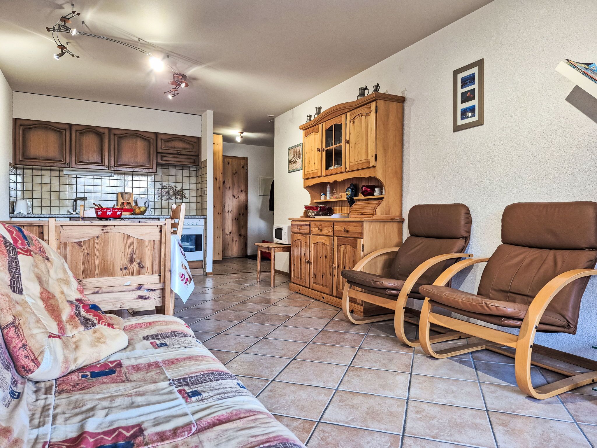 A cozy living room with a sofa, two brown leather chairs, a wooden cabinet, and a kitchen area with wooden cabinets and a tiled backsplash.