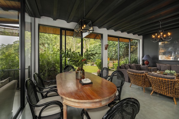 A dining area with a wooden table, black chairs, wicker sofas, and a large window overlooking greenery.