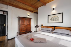 A bedroom with a wooden ceiling, a large wardrobe, a bed with grey linens, a framed artwork on the wall, and a nightstand with a lamp.