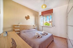 A bedroom with a bed, bedside tables, a desk, a hanging light fixture, a window with a view, and a wall-mounted decorative item.