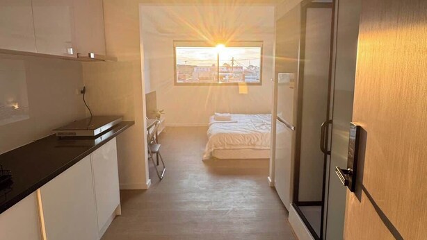 Een smalle, goed verlichte gang met een bed, een stoel en een kitchenette.