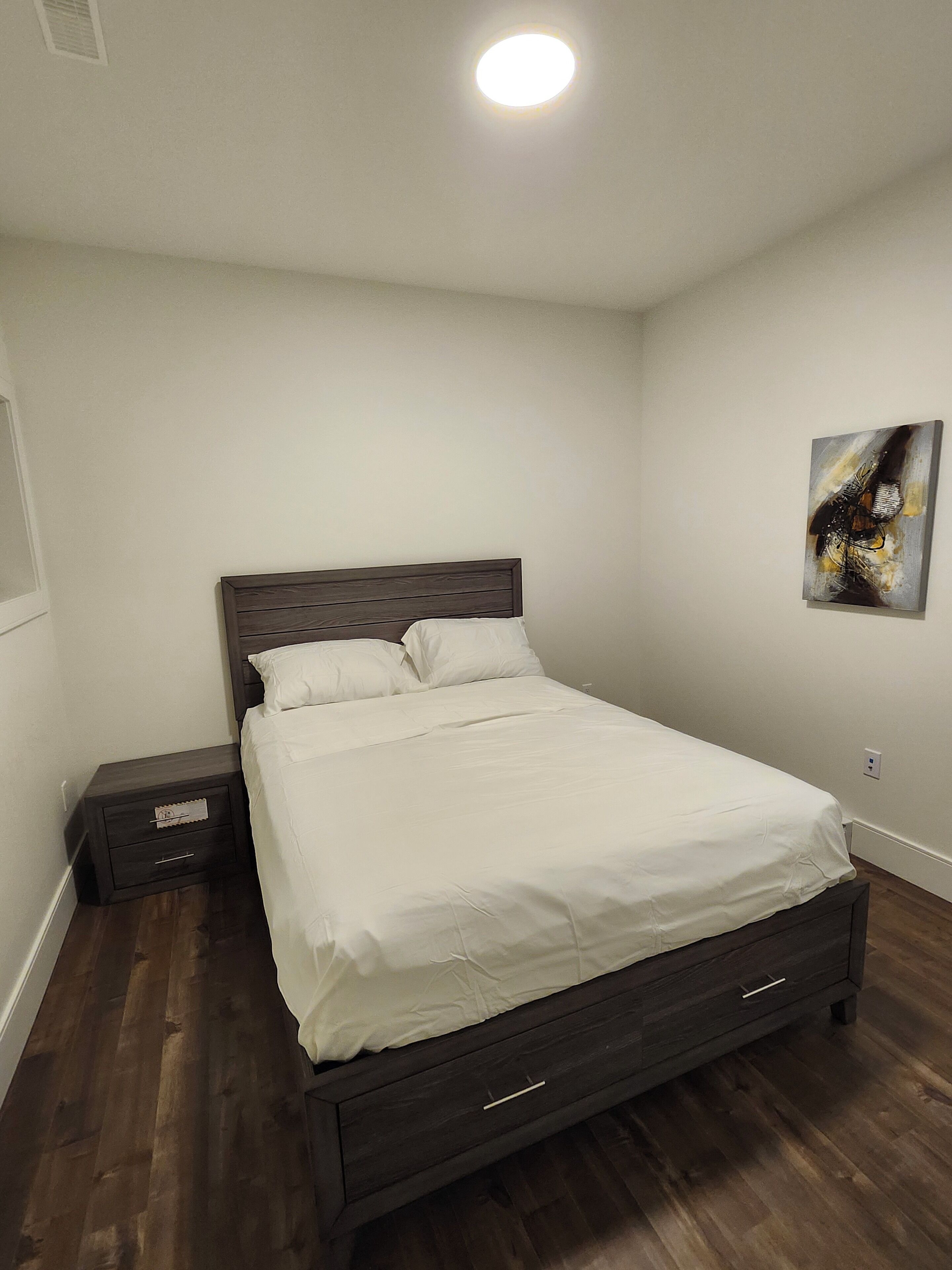 A neatly made bed in a room with a dark wooden headboard, a small bedside table, and a wall-mounted artwork.