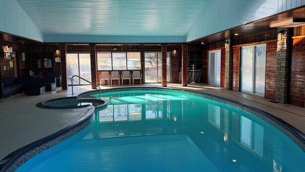 Una piscina interior con terraza de madera, zona de estar y grandes ventanales.