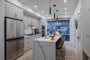 A modern kitchen with stainless steel appliances, a marble island, and a view through large windows. - Golf Course, Fireplace, Garage, Rooftop Patio (Calgary)