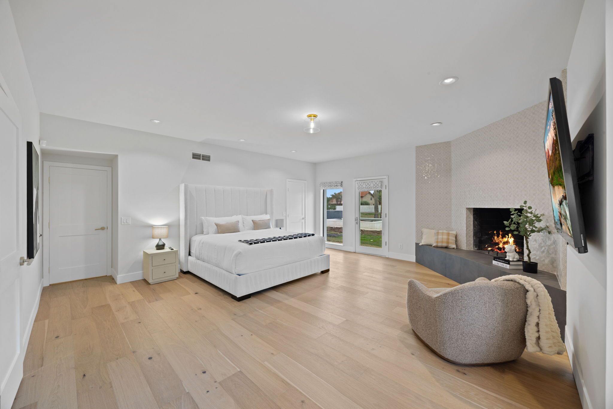 A modern bedroom with a large bed, a bedside table with a lamp, a fireplace, and a view of the outdoors.