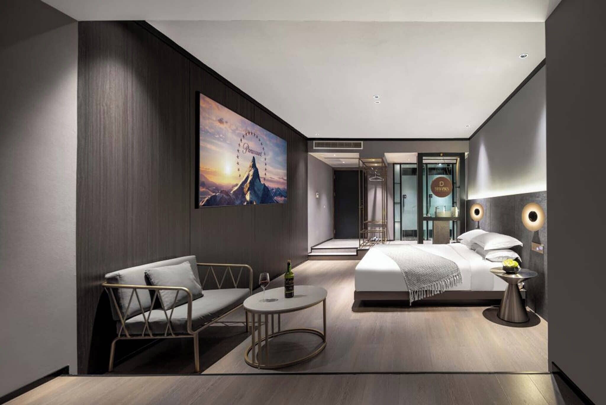 Une chambre d’hôtel moderne, équipée d’un lit, d’un coin salon avec un canapé et une petite table, d’un grand tableau au mur et offrant une vue sur le paysage urbain.
