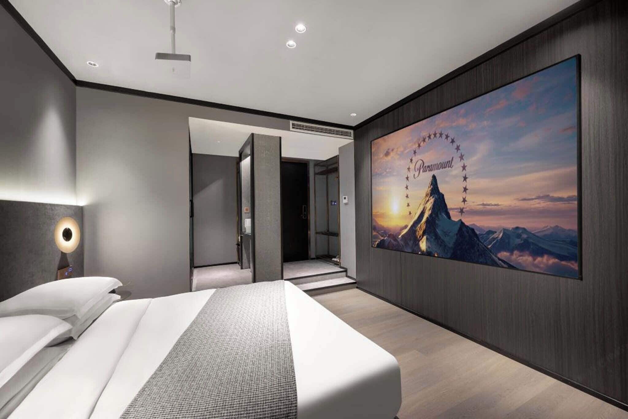 A modern hotel room with a large bed, a nightstand, a wall-mounted TV, and a view of a mountain range.
