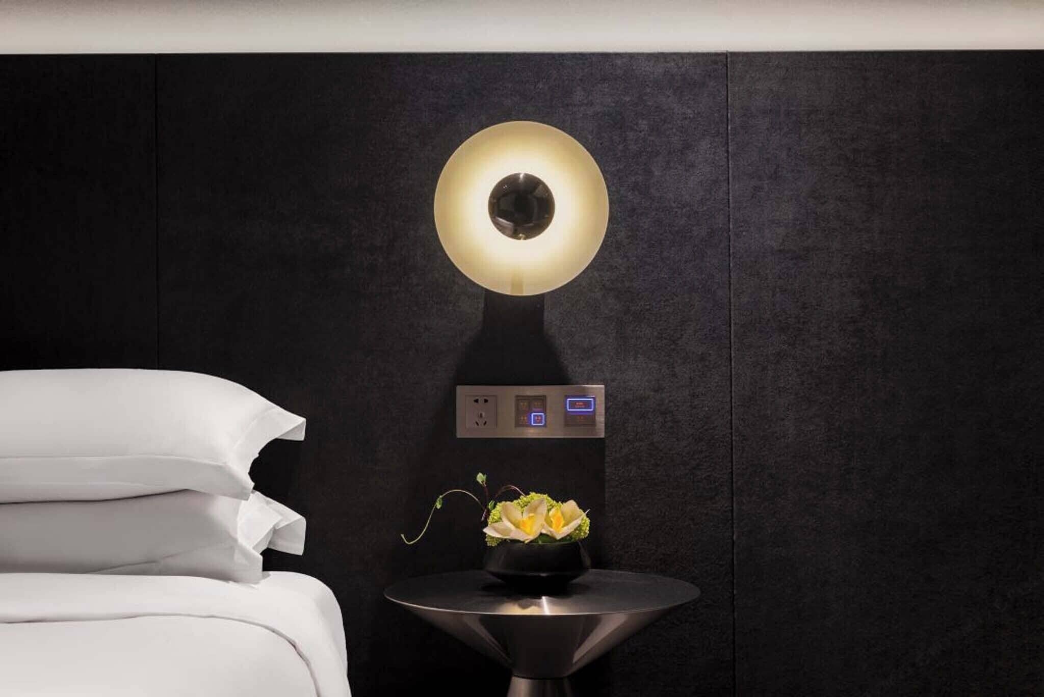 Habitación de hotel moderna con una cama, una mesita de noche con un florero y una lámpara fijada a la pared.