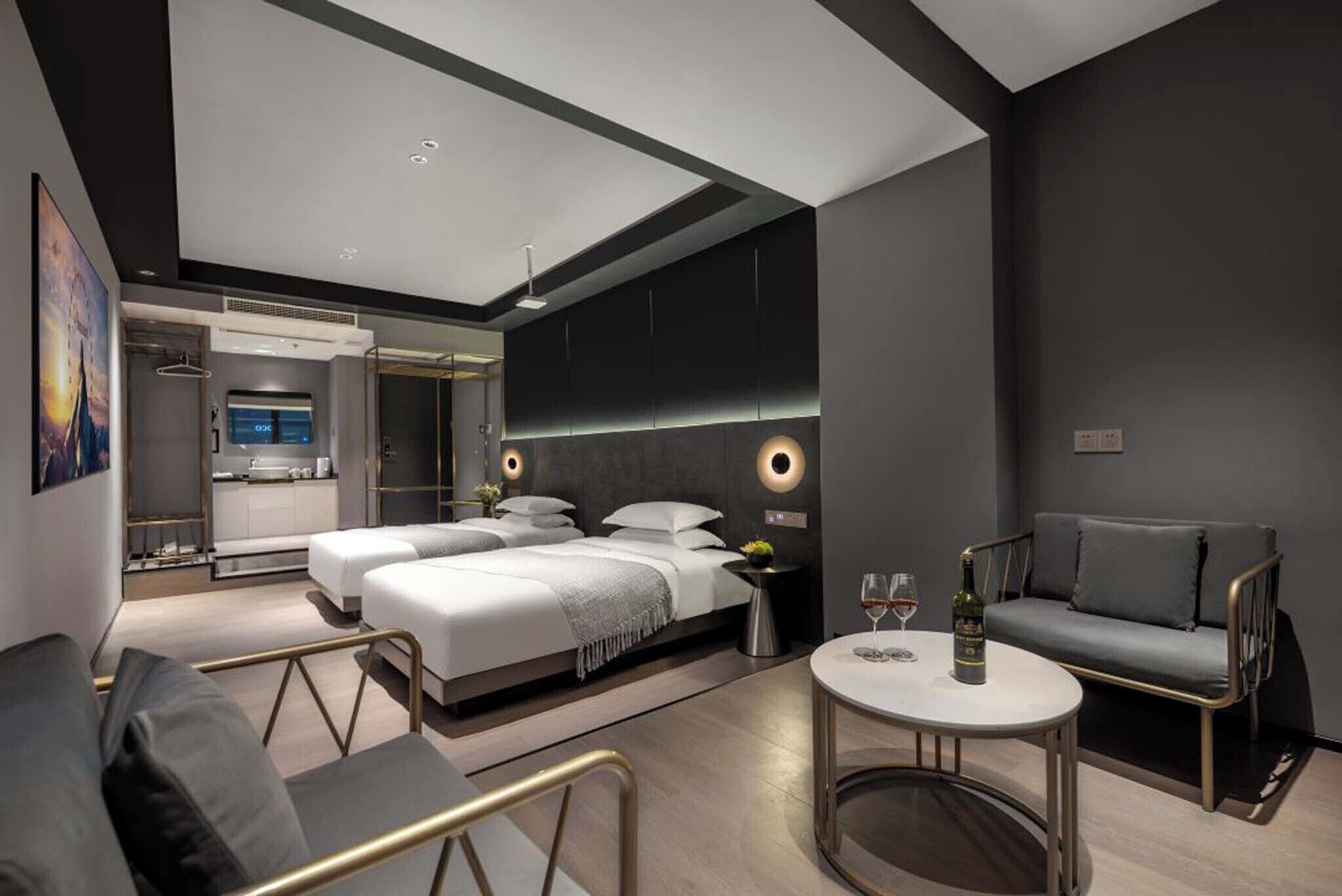 A modern hotel room with a large bed, a sofa, a small table with a bottle and glasses, and a kitchen area visible through an open door.