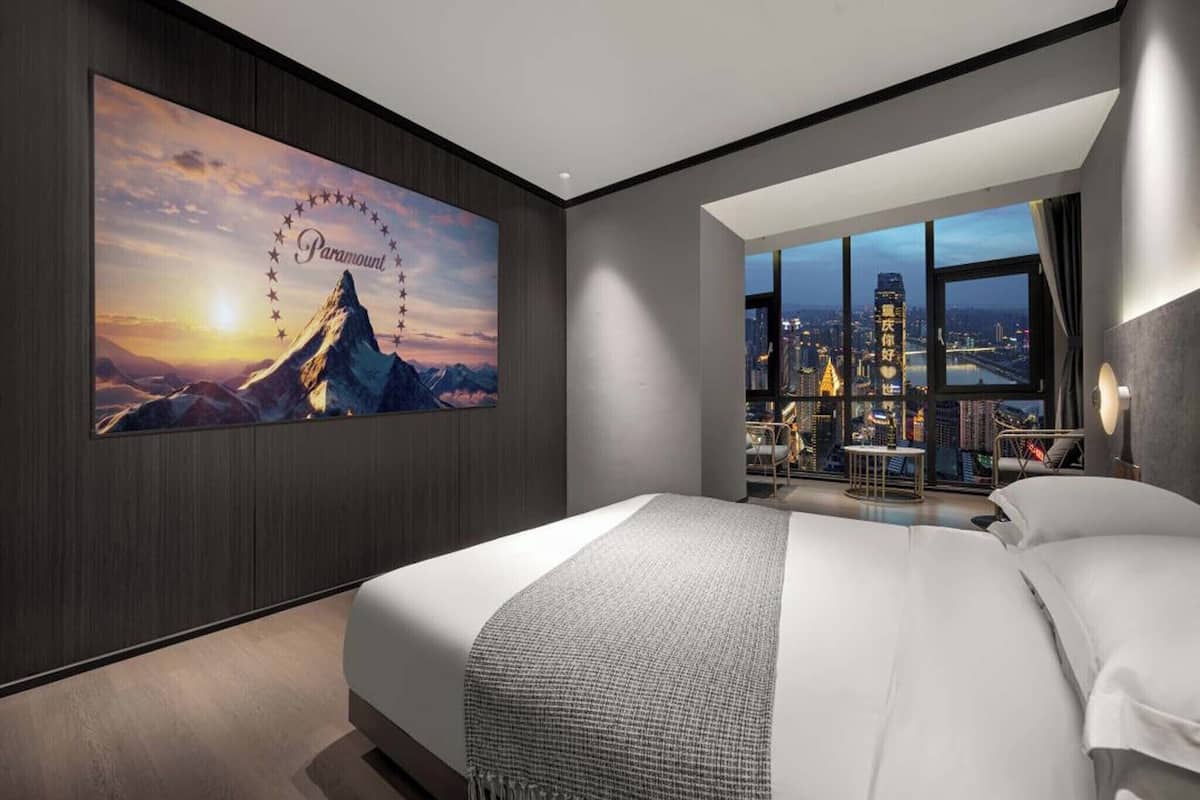 Une chambre d’hôtel moderne avec un grand lit, une vue sur la ville à travers de grandes fenêtres et une affiche de film Paramount accrochée au mur.