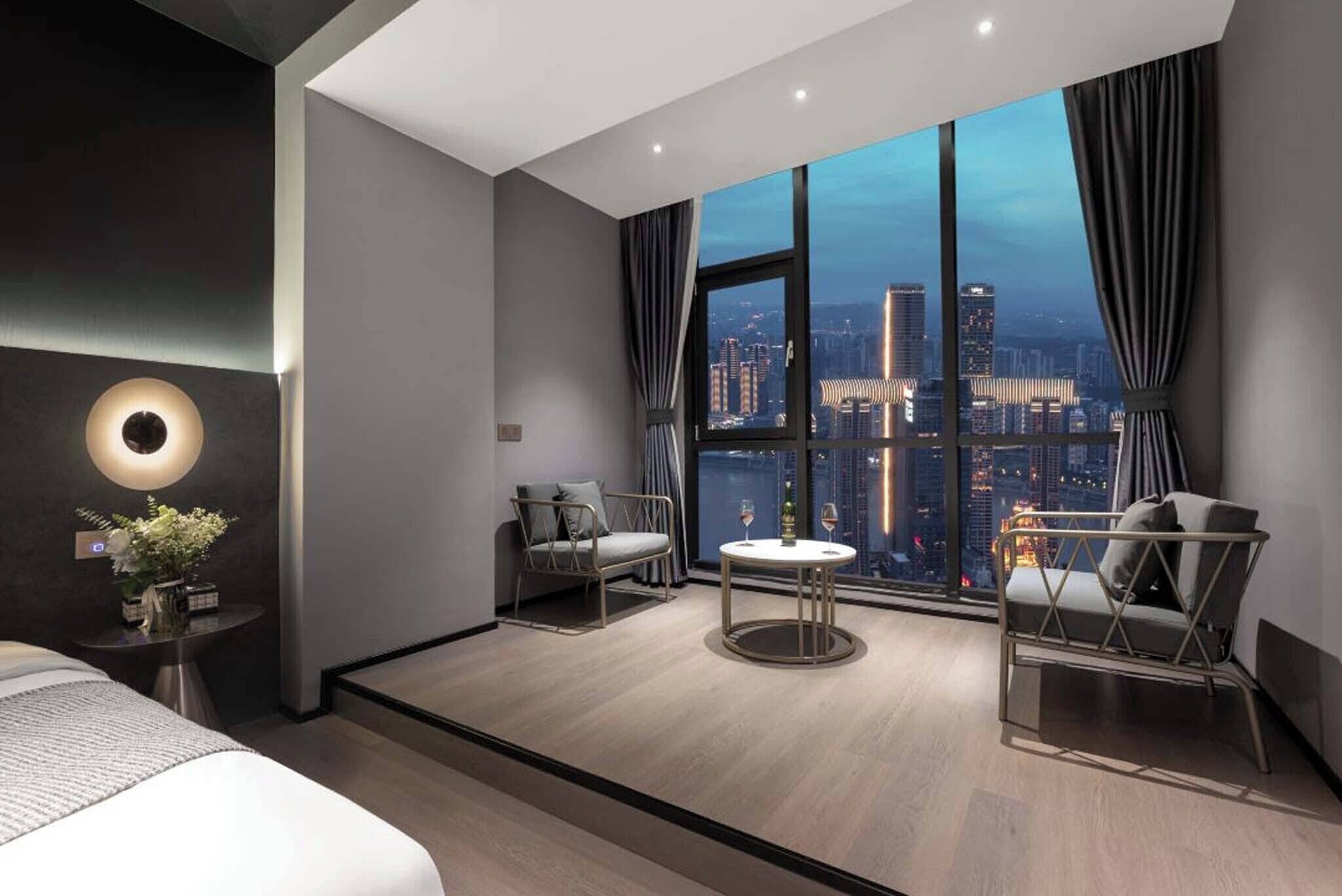 A modern hotel room with a large city view, a round table, two armchairs, and a bed.