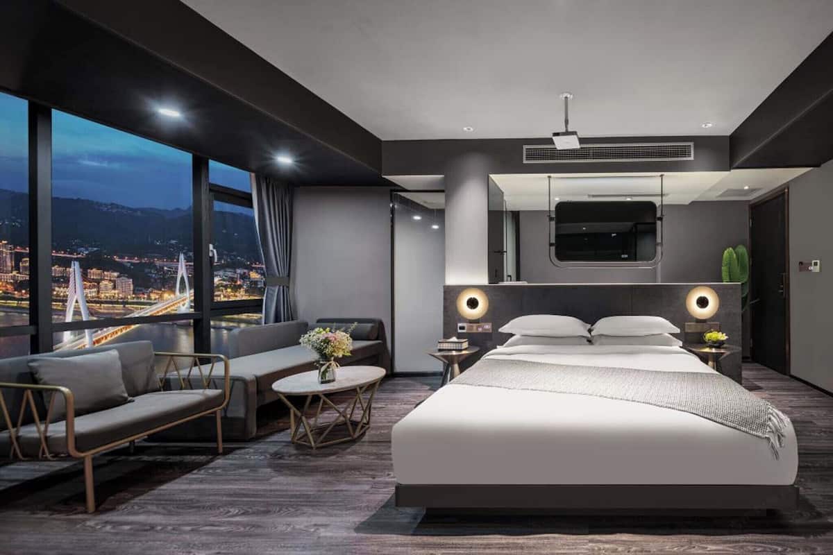 Une chambre d’hôtel moderne dotée d’un grand lit, d’un canapé, d’une petite table et offrant une vue sur le paysage urbain grâce à de grandes fenêtres.