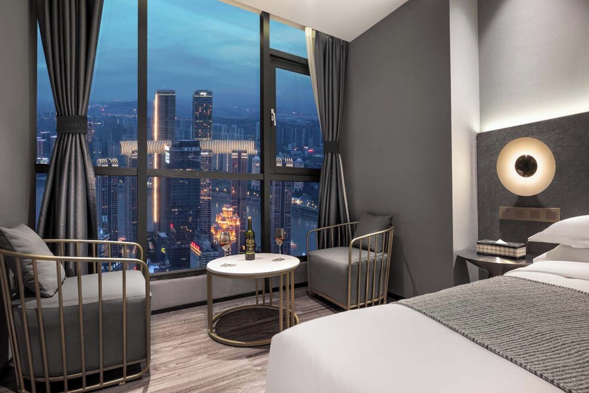 A modern hotel room with a large window offering a city view, a round table with a bottle and glasses, a bed, and a chair.