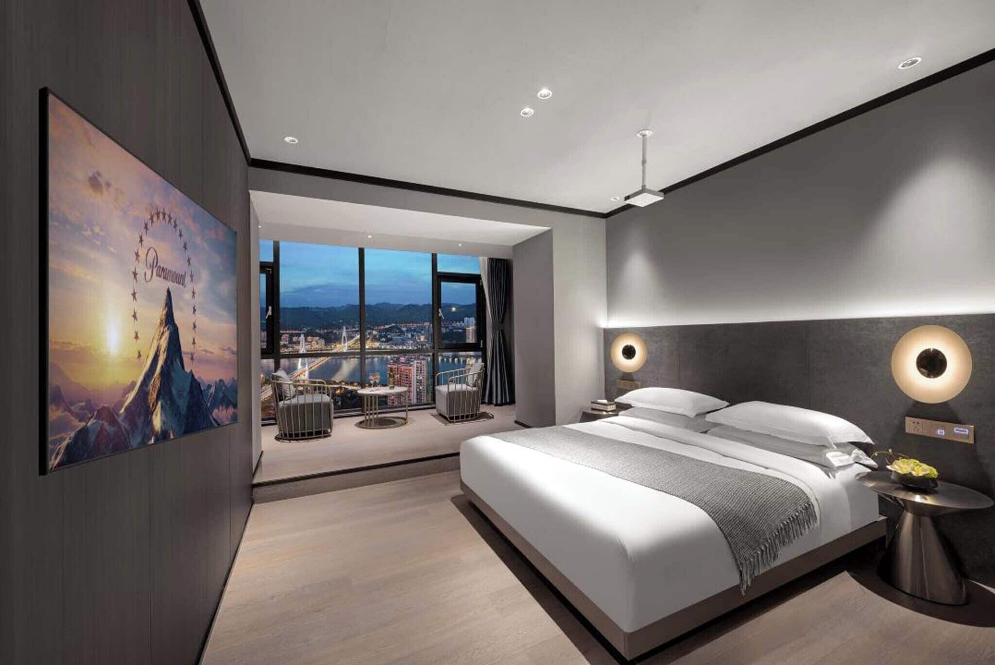 A modern hotel room with a large bed, a view of the city, and a balcony with outdoor seating.