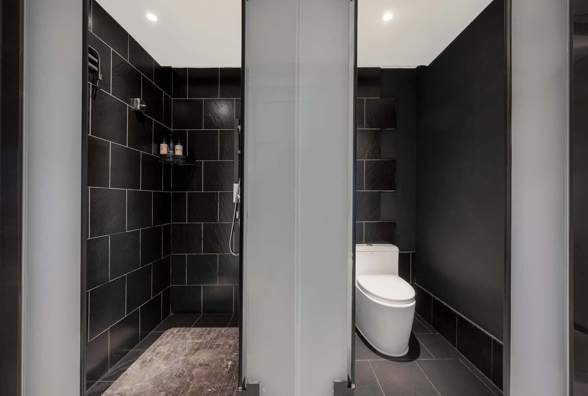 Un baño moderno con azulejos negros, cabina de ducha de vidrio y un inodoro blanco.