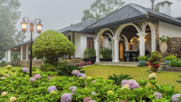 Una casa con porche cubierto, columnas de piedra y un jardín bien cuidado con hortensias.