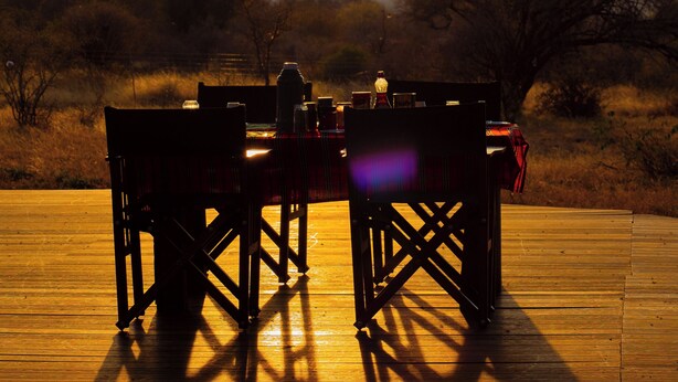 A wooden deck with a table and chairs, set against a backdrop of trees and a sunset. - Shibam Campsite (Naberera)