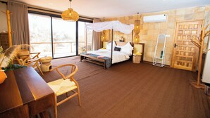 A spacious bedroom with a large bed, a wooden desk, wicker chairs, and a view of the outdoors. - Shibam Campsite (Naberera)
