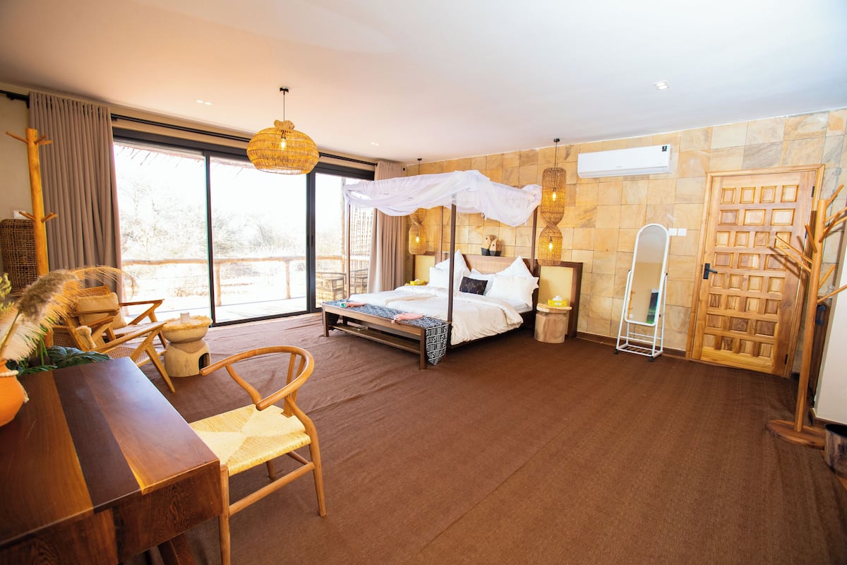 Une chambre spacieuse avec un grand lit, un bureau en bois, des chaises en osier et une vue sur l’extérieur.